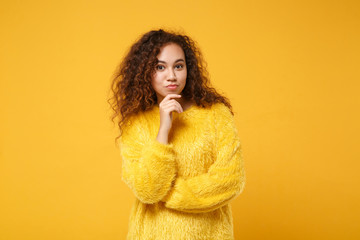 Pensive young african american girl in fur sweater posing isolated on yellow orange background studio portrait. People sincere emotions lifestyle concept. Mock up copy space. Put hand prop up on chin.