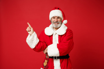 Fototapeta na wymiar Smiling elderly gray-haired bearded mustache Santa man in Christmas hat posing isolated on red background. New Year 2020 celebration holiday concept. Mock up copy space. Pointing index finger aside.