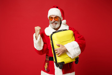 Elderly gray-haired mustache bearded Santa man in Christmas hat sunglasses posing isolated on red background. New Year 2020 celebration concept. Mock up copy space. Hold suitcase doing winner gesture.