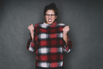 Angry young woman in glasses grinning with eyes closed and raised hands