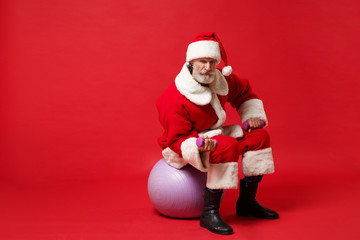 Elderly gray-haired mustache bearded Santa man in Christmas hat posing isolated on red background. Happy New Year 2020 celebration holiday concept. Mock up copy space. Sit on fitball, hold dumbbells.