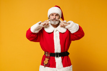 Fototapeta na wymiar Elderly gray-haired mustache bearded Santa man in Christmas hat isolated on yellow background. Happy New Year 2020 celebration holiday concept. Mock up copy space. Pointing fingers on blowing cheeks.