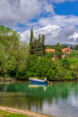 Beautiful landscape with pond, blue fisher boat, reflecting on calm water surface, green foliage of trees and white clouds on blue sky. Corfu Island, Greece.