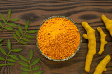 Turmeric powder in glass bowl and turmeric roots on wooden background with green leaves.