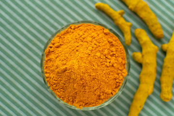 Turmeric powder in glass bowl and turmeric roots at green pastel kitchen towel background. Indian spices. Trendy healthy food.