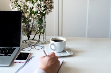 Home office desk in white colors with woman hand writing on a notebook with a pen with laptop, cup of coffee, notebook, phone, glasses on a white background. Business womans workplace and objects.