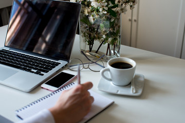 Home office desk in white colors with woman hand writing on a notebook with a pen with laptop, cup of coffee, notebook, phone, glasses on a white background. Business womans workplace and objects.