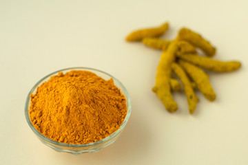 Blurred turmeric powder in glass bowl with dry roots or rhizoms at ivory pastel background. Southeast asia spices.