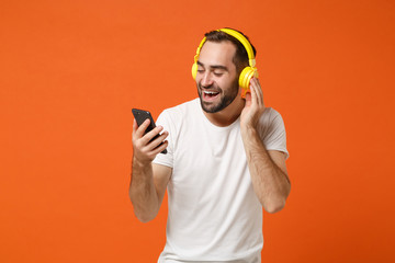 Cheerful young man in casual white t-shirt posing isolated on orange wall background studio portrait. People lifestyle concept. Mock up copy space. Listening music with headphones, hold mobile phone.