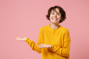 Smiling joyful young brunette woman girl in yellow sweater posing isolated on pink wall background studio portrait. People sincere emotions lifestyle concept. Mock up copy space. Pointing hands aside.