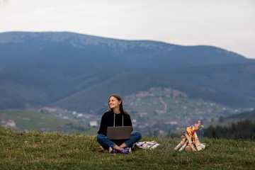 Fototapeta na wymiar young woman freelancer with laptop in the mountains in the evening. Tourist girl sitting near campfire.