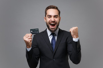 Happy young business man in classic black suit shirt tie posing isolated on grey background. Achievement career wealth business concept. Mock up copy space. Hold credit bank card doing winner gesture.
