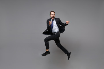 Fototapeta na wymiar Cheerful young business man in classic black suit shirt tie posing isolated on grey background. Achievement career wealth business concept. Mock up copy space. Jumping, point index fingers on camera.