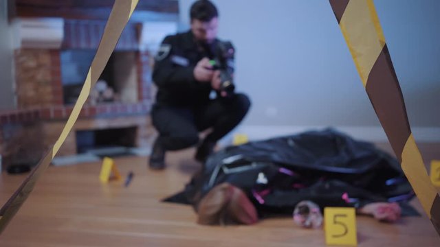 Blurred Caucasian police officer taking photos of covered murdered body and evidences standing around. Focused on police tape at the foreground. Detective, investigation, crime scene.