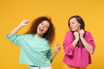 Two funny women friends european african american girls in pink green clothes posing isolated on yellow background. People lifestyle concept. Mock up copy space. Have fun dancing keeping eyes closed.