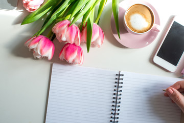 Young woman hands hold opened notebook pages. Morning coffee mug for breakfast, empty notebook, pencil and pink tulips flowers on white table top view in flat lay style. Woman working desk.
