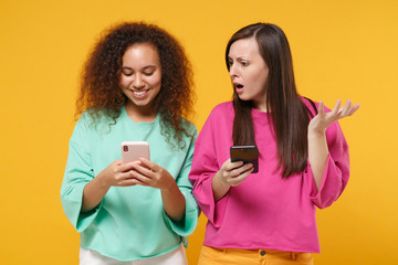 Two shocked women friends european african american girls in pink green clothes posing isolated on yellow background. People lifestyle concept. Mock up copy space. Using mobile phone, typing message.