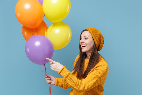 Cute young woman in sweater hat posing isolated on blue background. Birthday holiday party people emotions concept. Mock up copy space. Celebrating hold colorful air balloons point index finger aside.