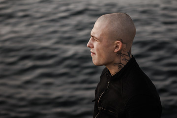 A stylish guy in tattoos stands on the seashore on the breakwaters and looks thoughtfully into the distance. Brutal brutal young man with a tattoo on his neck outdoors. Stylish and fashionable guy.