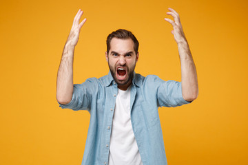 Fototapeta premium Angry young bearded man in casual blue shirt posing isolated on yellow orange wall background studio portrait. People emotions lifestyle concept. Mock up copy space. Screaming, spreading rising hands.