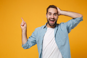 Excited young man in casual blue shirt posing isolated on yellow orange wall background, studio portrait. People lifestyle concept. Mock up copy space. Pointing index finger up, putting hand on head.