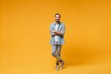 Fototapeta na wymiar Laughing young bearded man in casual blue shirt posing isolated on yellow orange wall background, studio portrait. People sincere emotions lifestyle concept. Mock up copy space. Holding hands crossed.