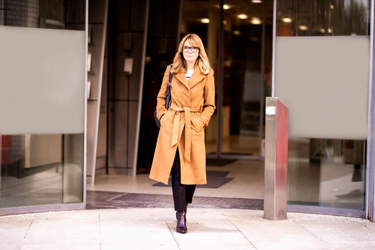  Shot Of Middle Aged Woman In Beige Coat With A Bag Walking Out Of The Office Building