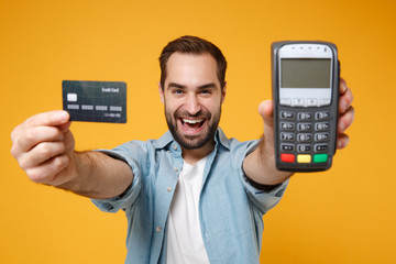 Cheerful young man in blue shirt posing isolated on yellow orange background. People lifestyle concept. Mock up copy space. Hold wireless bank payment terminal to process acquire credit card payments.