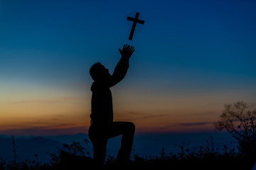 Silhouette of a young Christian man who is praying on the holy cell in the Bible and raising a cross over a Christian with a light sunset background. Christian concept