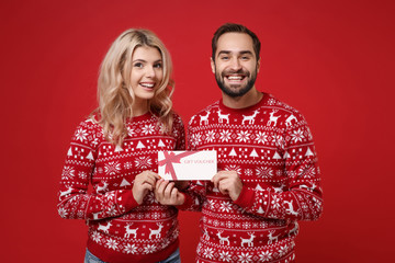 Smiling young couple guy girl in Christmas knitted sweaters posing isolated on bright red...