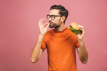 Young man holding a piece of hamburger. Student eats fast food. Burger is not helpful food. Very hungry guy. Diet concept. Isolated over pink background.