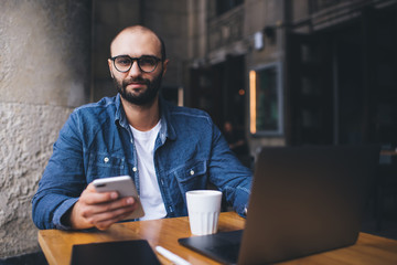 Attentive modern man in glasses texting on smartphone while resting at street cafe