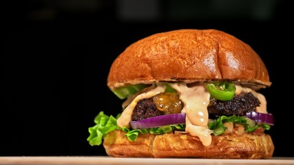 Craft burger is cooking on black background. Consist: red sauce salsa, lettuce, red onion, pickle, cheese, chilli green pepper, air bun and marbled meat beef. Not made ideal. Loving hand made.
