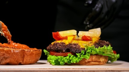Craft burger is cooking on black background in black food gloves. Consist: red sauce salsa, lettuce, red onion, pickle, cheese, chilli green pepper, air bun and marbled meat beef. Not made ideal