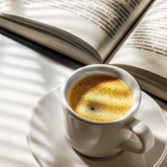a Cup of black coffee and an open book on a white table and the shade from the blinds