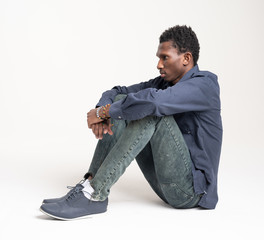 A black guy (man) in a blue shirt and jeans is sitting on a white background