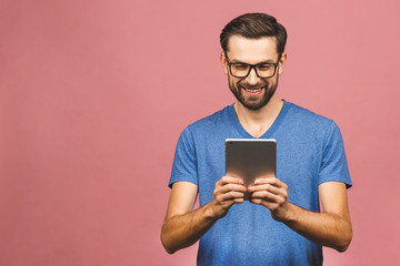 Happy young man in casual standing and using tablet isolated over pink background.