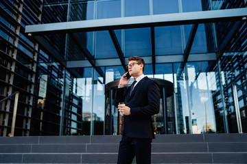 Fototapeta premium Trendy formal man speaking on mobile phone Young confident businessman in formal suit holding disposable cup of coffee while talking on smartphone standing against steps of office building