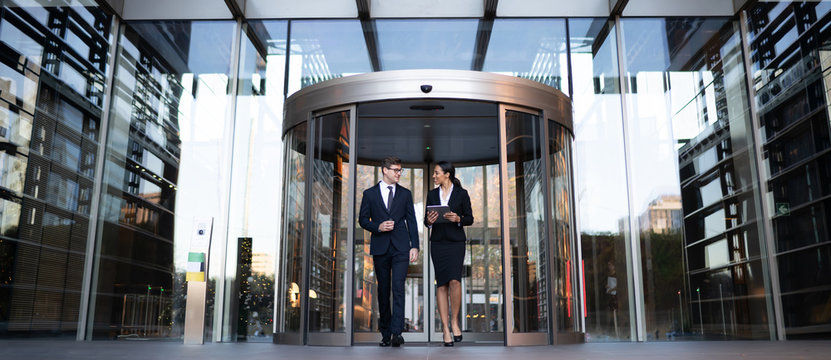 Smiling Young Coworkers Talking On Street
Contemporary Elegant Businesswoman With Tablet Talking To Male Colleague While Walking Out Of Modern Office Building With Glass Revolving Doors