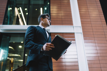 Fototapeta premium Contemporary formal man with paper file From below of elegant man in glasses holding file with papers while standing on background of contemporary city building looking away