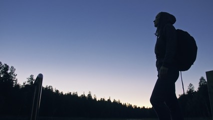 Silhouette traveler photographing scenic view in forest, river. Wood pier. One woman shooting nice dark magic night look. Girl take photo video on camera. Photographer walk with backpack. Outdoor.