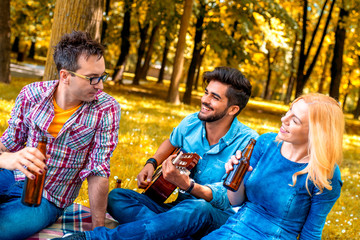 Friends in autumn park enjoying time together, playing guitar and drinking beer