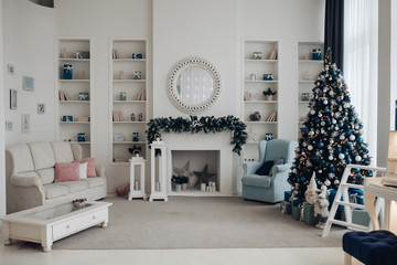 View over white modern room decorated for Christmas holidays. Decorated and illuminated Christmas tree, fireplace with fir branch, candles and handmade paper stars in fireplace.