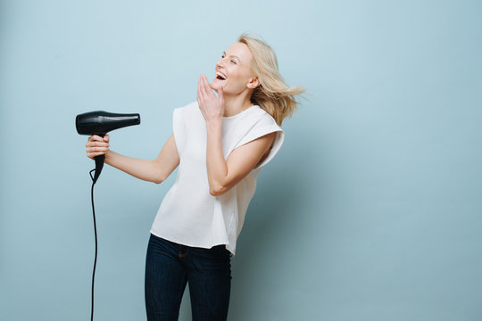 Playful fooling blonde woman in a white shirt is blowing herself with a hairdryer over blue background. She's laughing and amused, covering mouth with a hand.
