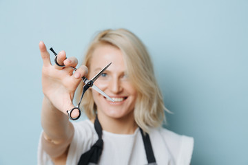 Playful smirking stylish blonde female hairdresser in apron holding open scissors in outstretched...