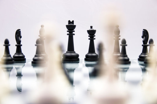 black chess pieces stand on a mirrored chess board with blurry white pieces in the foreground on a white background