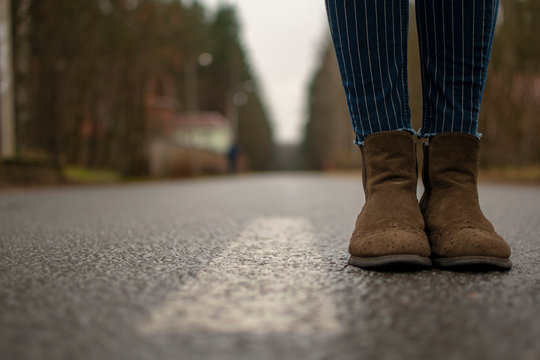 female legs in brown boots and jeans stand in the middle of the road near the markings. the road goes to the horizon. bottom view
