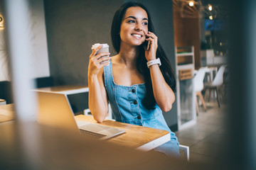 Happy female with coffee cup laughing during positive cellphone conversation with best friend, cheerful Ukrainian hipster girl using smartphone application for mobile phoning during break