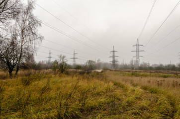 A transmission tower or electricity pylon. Steel lattice tower, used to support an overhead power line. The sky is covered with fog. Under the tower are trees and grass. Autumn landscape.