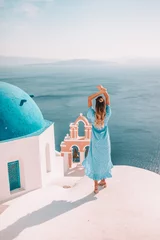 Zelfklevend Fotobehang Jonge vrouw met blond haar en blauwe jurk in oia, santorini, griekenland met uitzicht op de oceaan en kerken © Mathilda